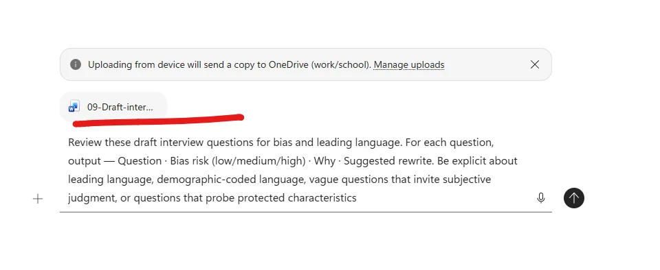 Microsoft 365 Copilot Chat composer with the file 09-Draft-interview attached as a Word file chip, underlined in red as an annotation. A system banner above reads Uploading from device will send a copy to OneDrive (work/school). Below the file chip the prompt is typed: Review these draft interview questions for bias and leading language. For each question, output — Question · Bias risk (low/medium/high) · Why · Suggested rewrite. Be explicit about leading language, demographic-coded language, vague questions that invite subjective judgment, or questions that probe protected characteristics. A plus upload button on the left, microphone and send arrow on the right of the composer.
